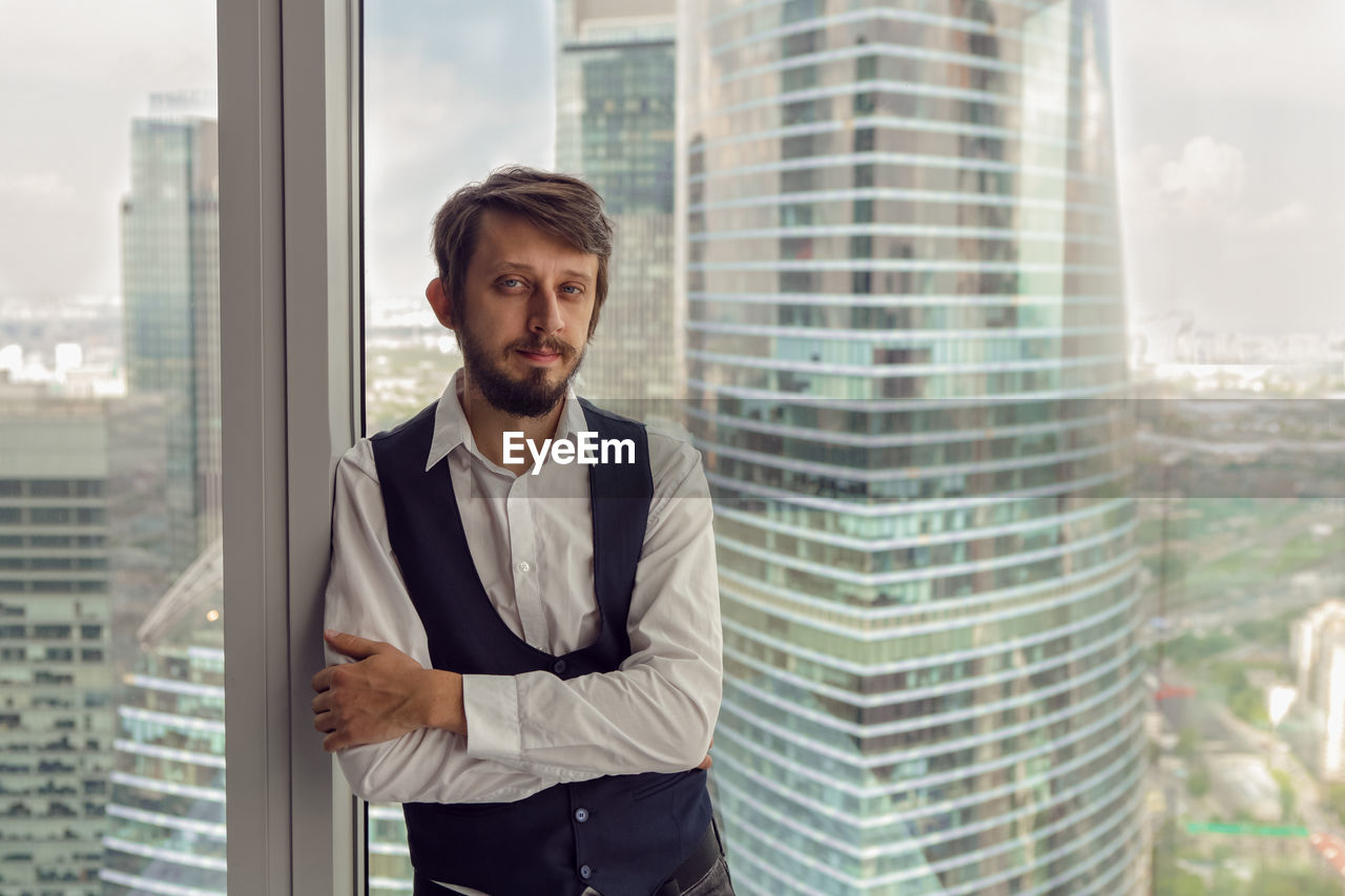 Thoughtful businessman with beard stands next to  window in office backdrop of skyscrapers in moscow