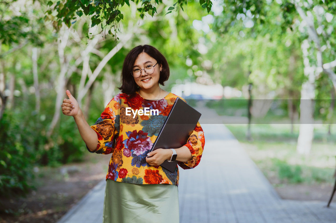 Smiling asian woman with glasses and a laptop showing thumbs up in park