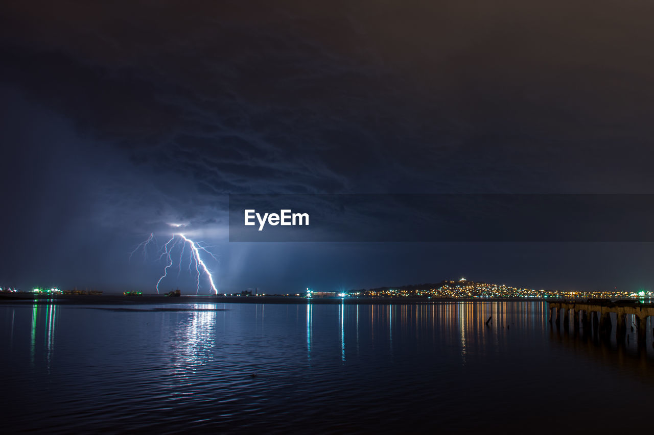 Illuminated city by sea against sky at night