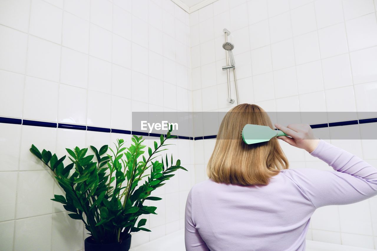 Rear view of woman holding hairbrush against wall in bathroom