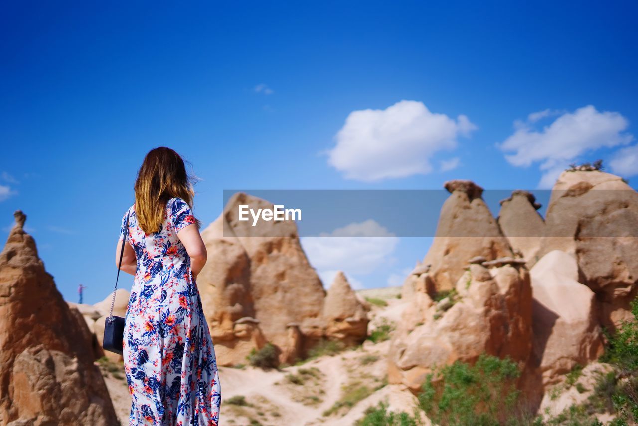 REAR VIEW OF WOMAN STANDING ON ROCK FORMATION AGAINST BLUE SKY