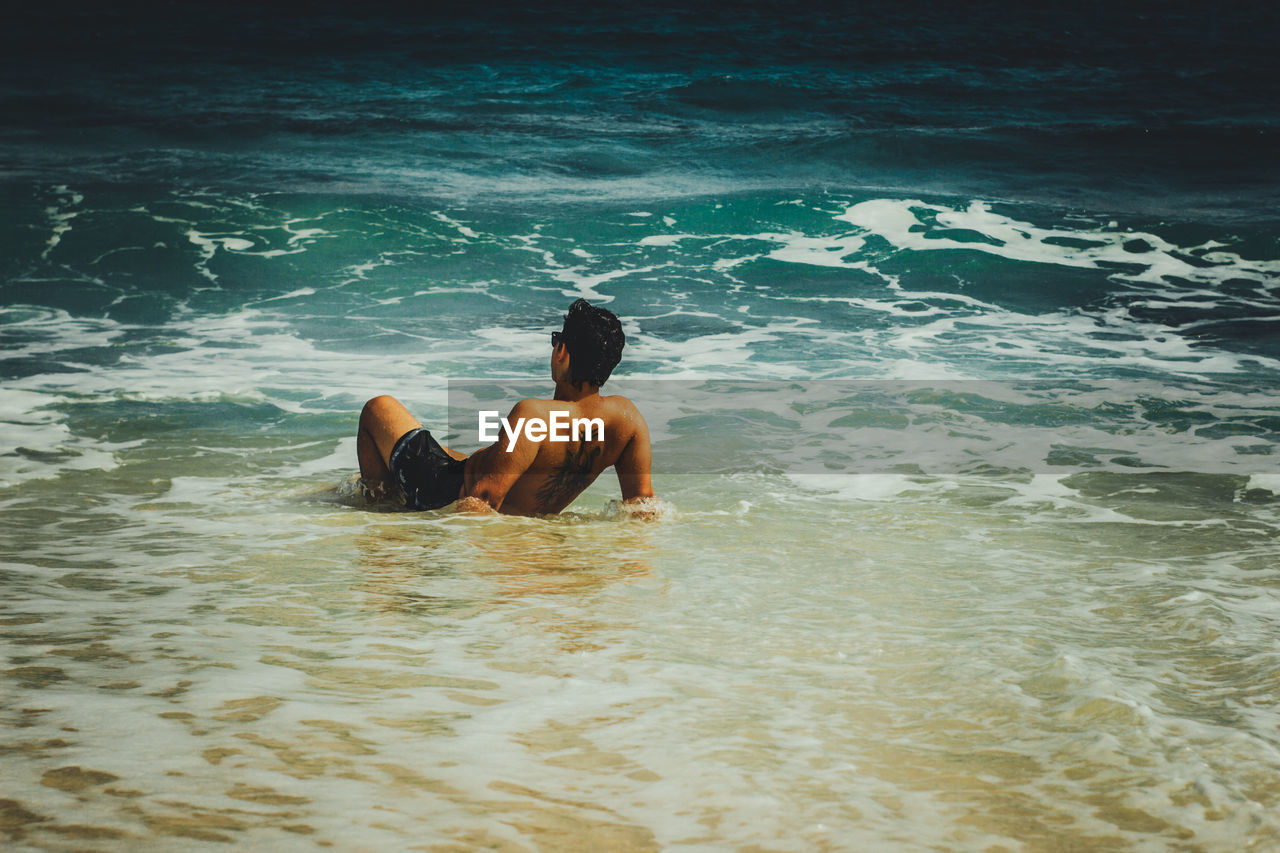 Rear view of shirtless man lying in sea