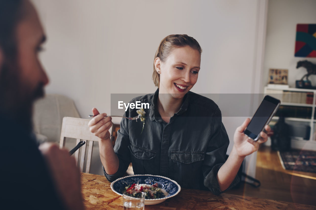 Happy woman showing mobile phone to man while eating pasta at home