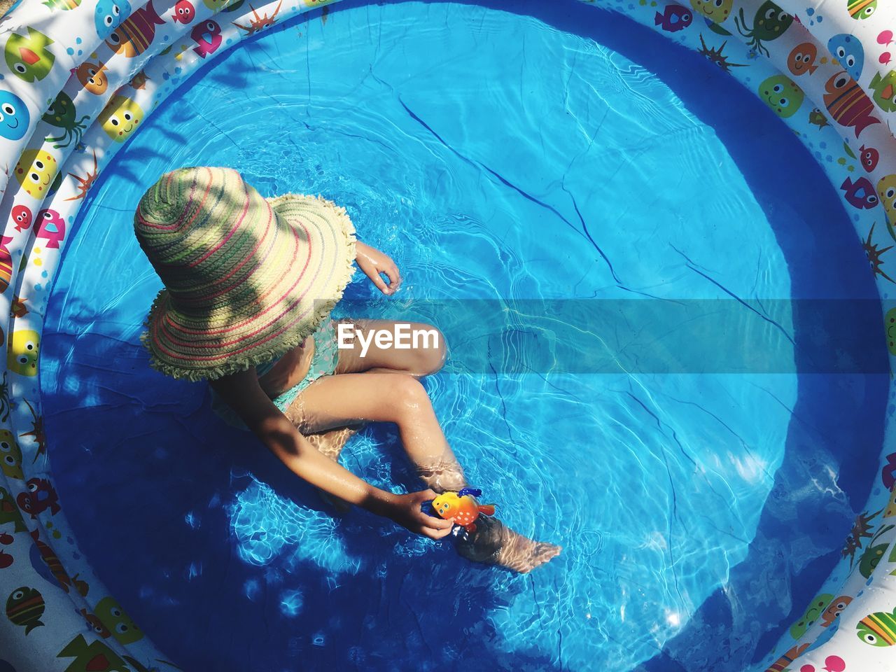 High angle view of girl wearing sun hat sitting in wading pool on sunny day