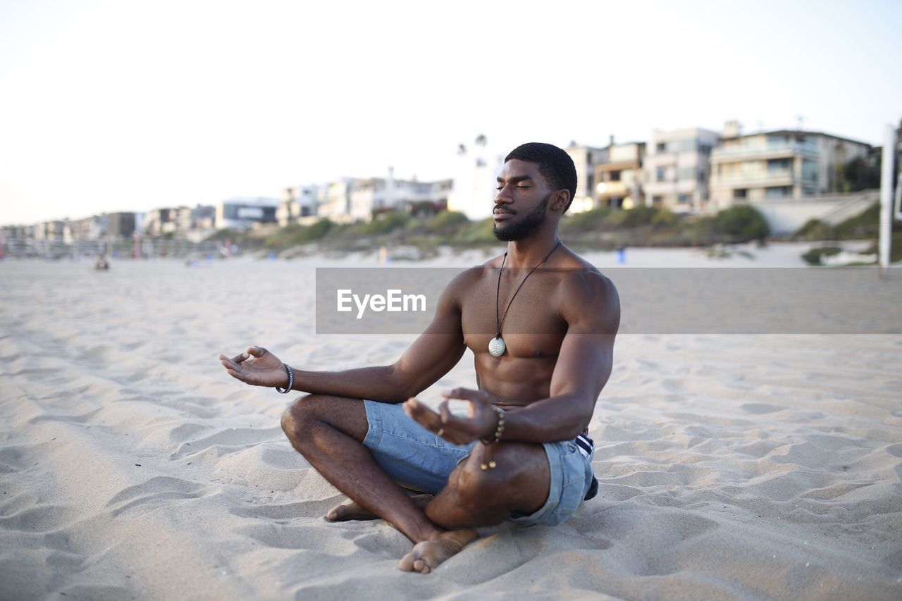 Shirtless muscular man meditating at beach against clear sky