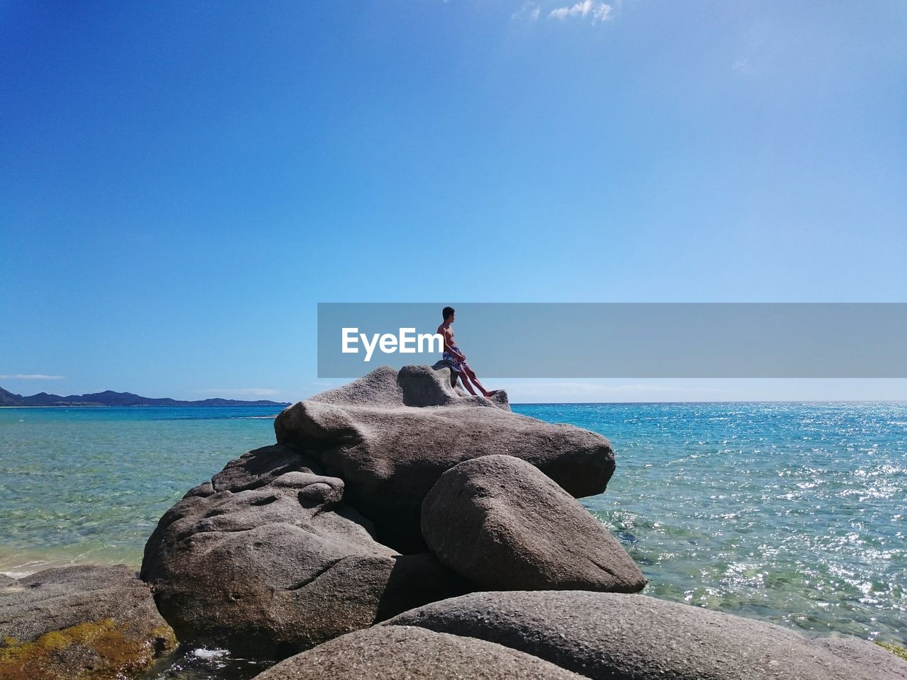 Man sitting on rocks at beach against blue sky