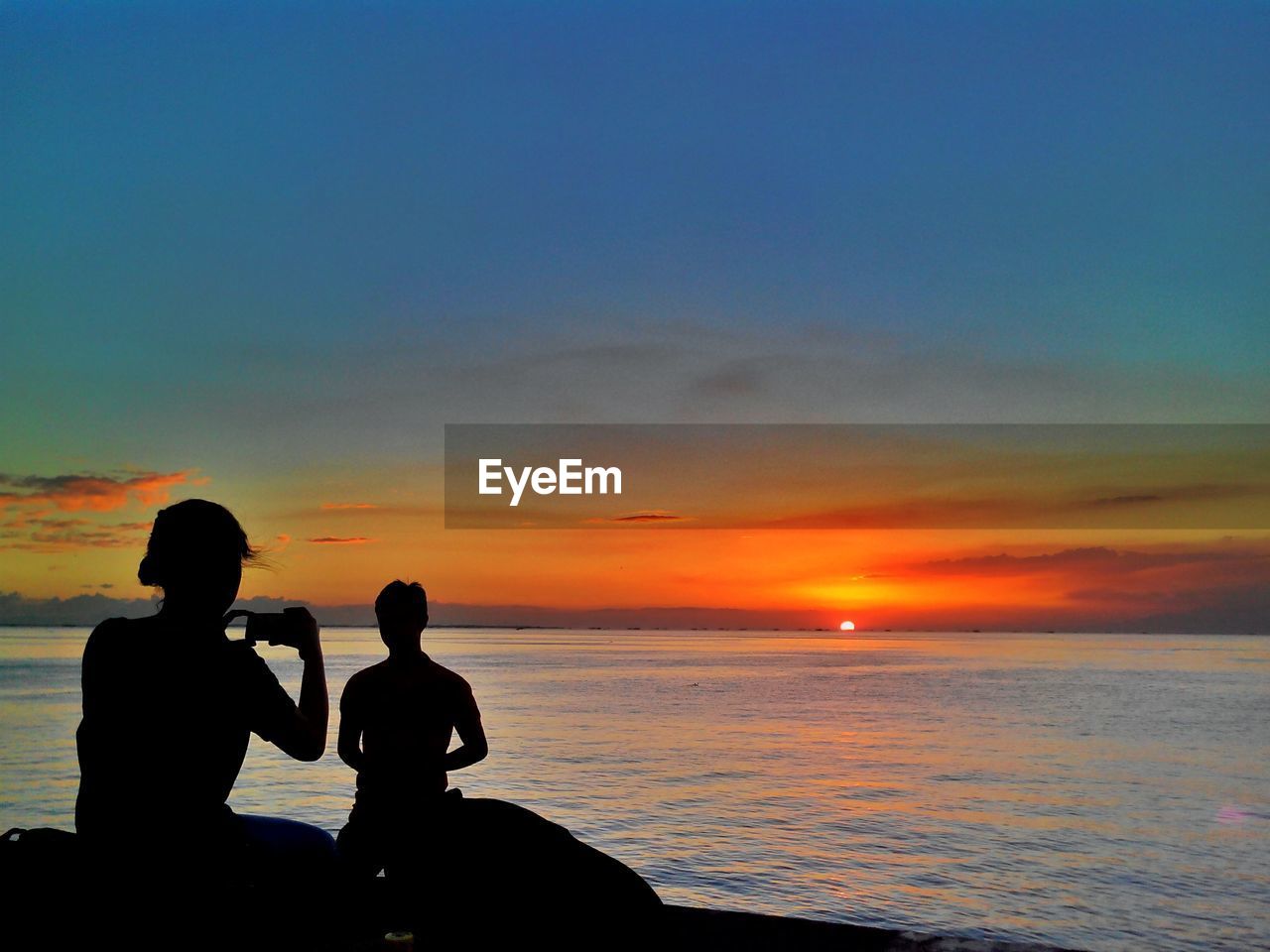 Silhouette woman photographing man by sea against sky during sunset