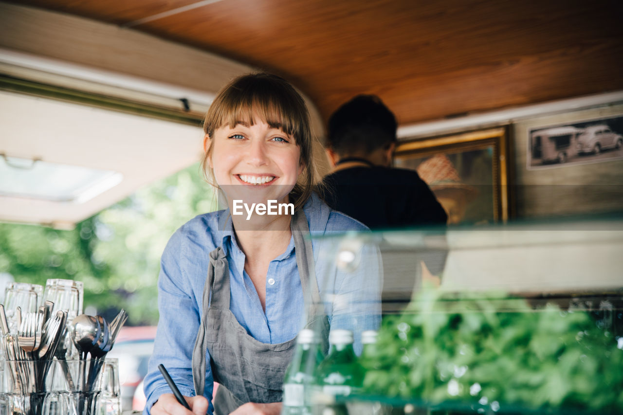 Portrait of happy female owner standing in food truck while coworker working in background
