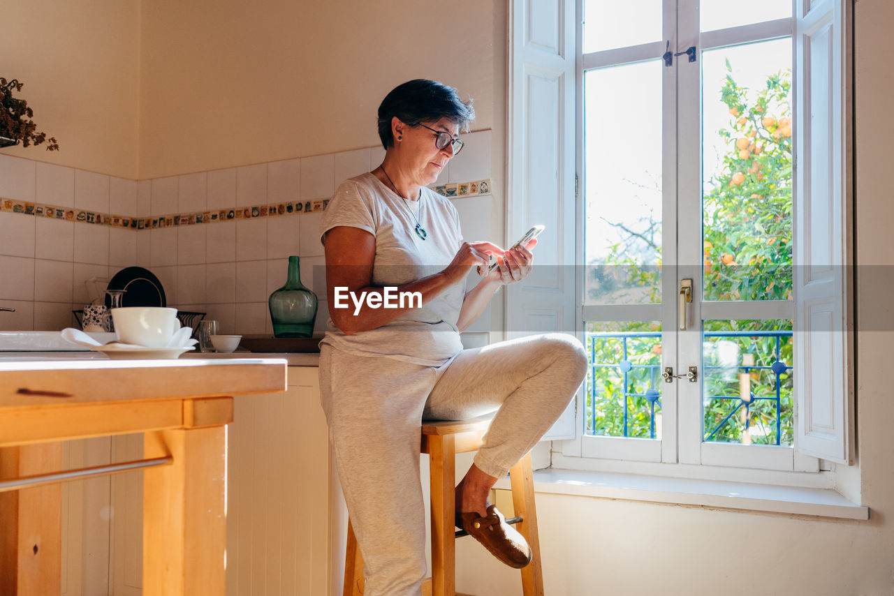 Mature female in casual clothes and glasses sitting on stool near window and browsing mobile phone in cozy home kitchen with wooden furniture in daylight