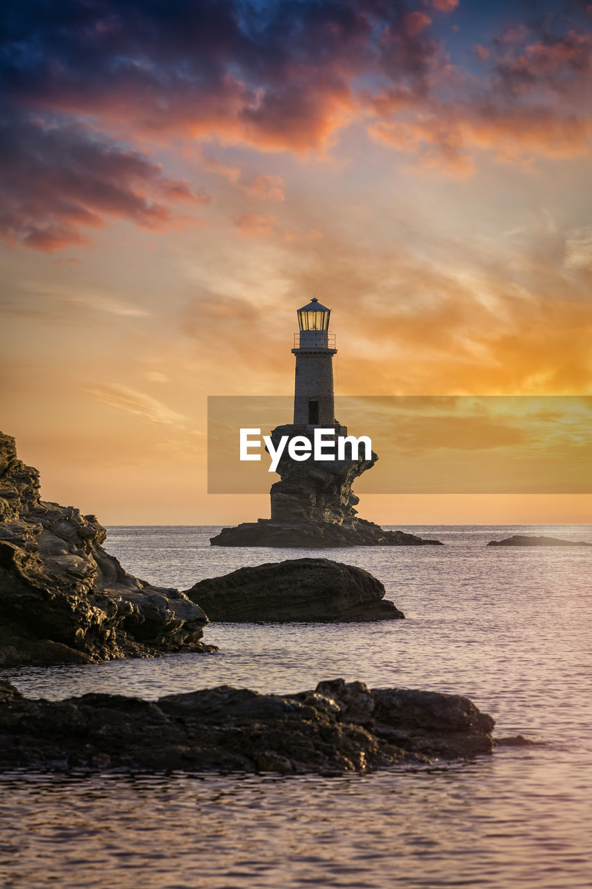 LIGHTHOUSE ON SEA BY BUILDING AGAINST SKY DURING SUNSET