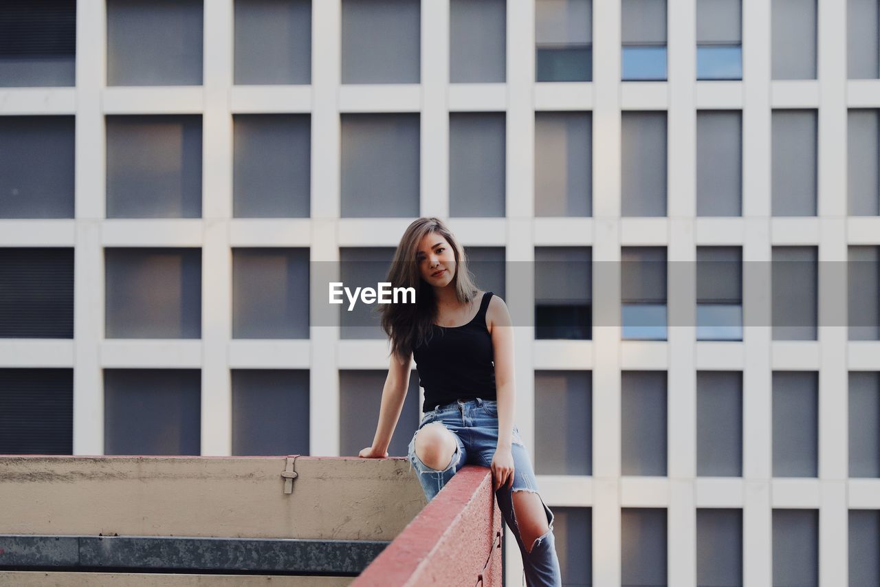 Portrait of young woman wearing torn jeans sitting on railing against modern building