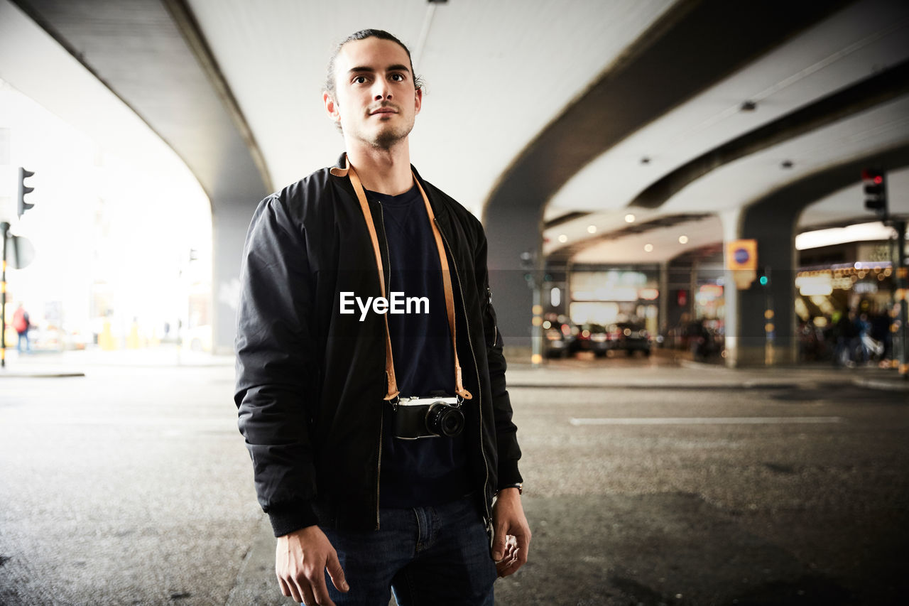 Young man with camera standing on street below overpass in city