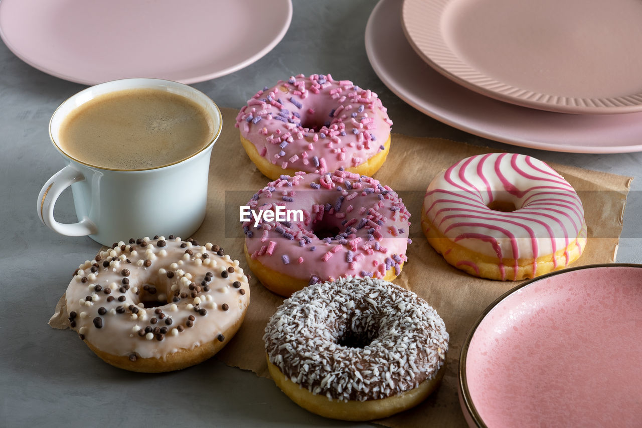 Donuts lie on craft paper. cup of coffee and pink plates stands next to donuts laid out on table. 