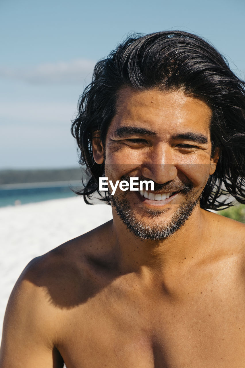 Close-up of happy shirtless man looking down while sitting at hyams beach against sky during sunny day