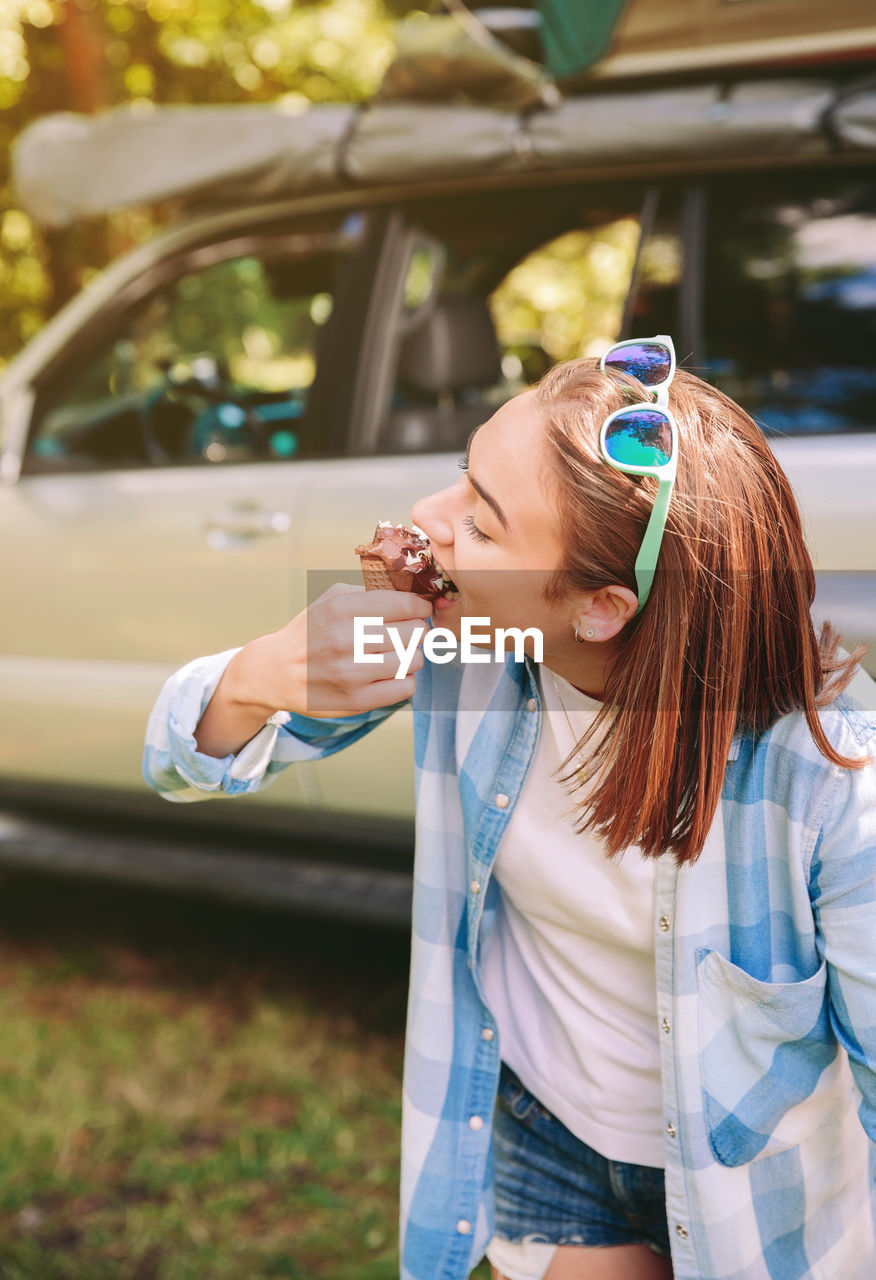 Young woman eating ice cream cone while standing against car