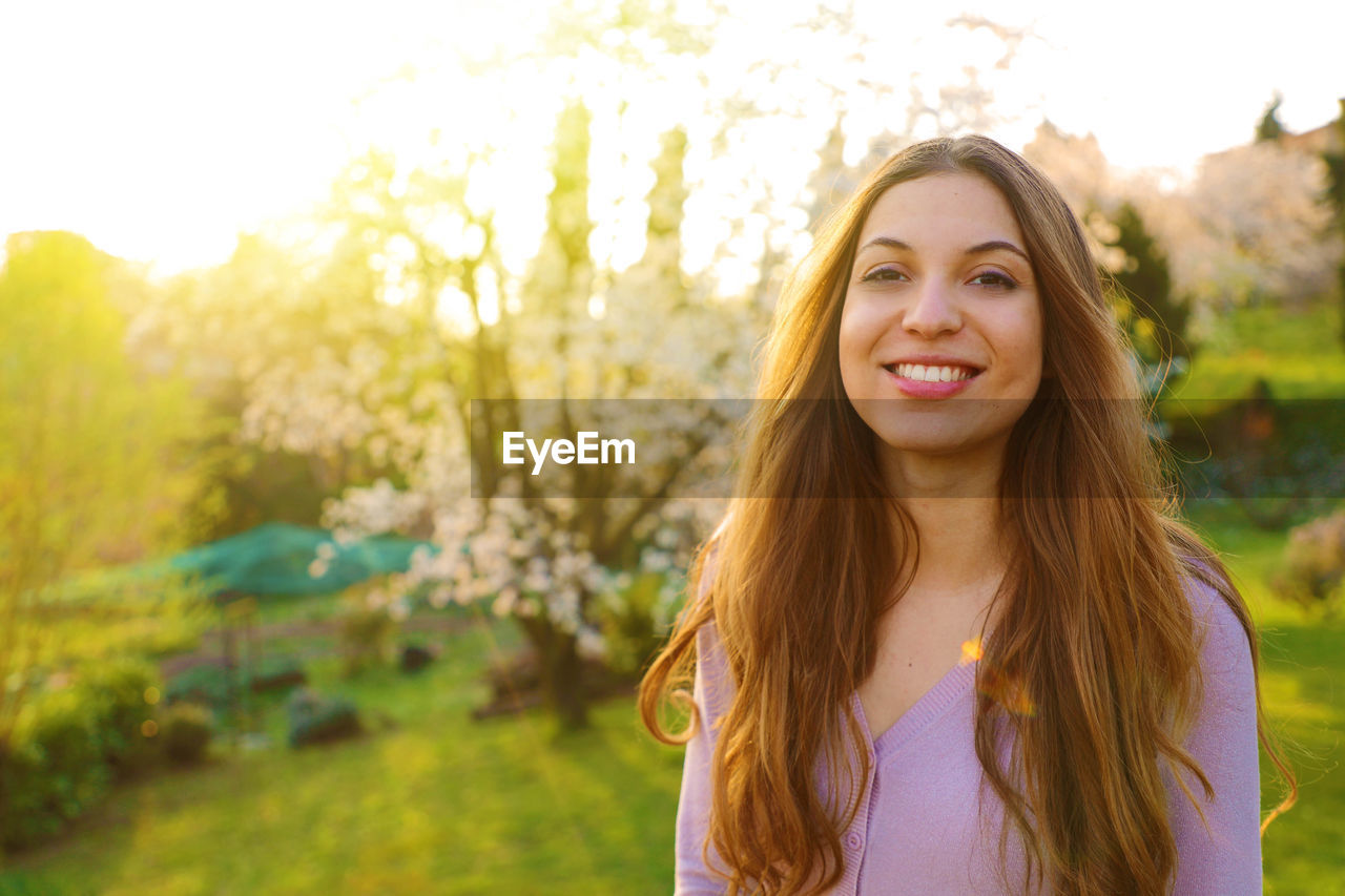 PORTRAIT OF A SMILING YOUNG WOMAN AGAINST TREES