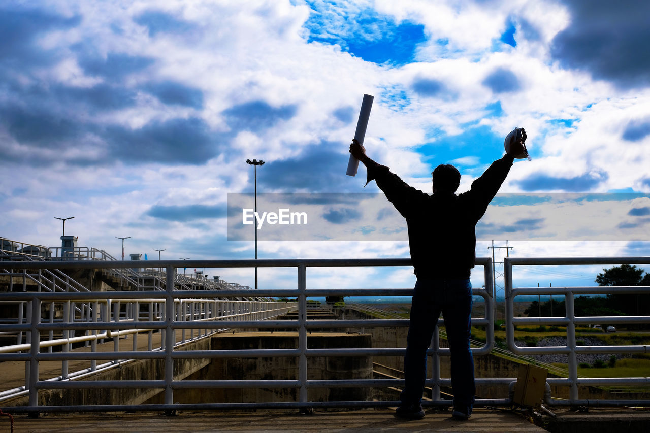 REAR VIEW OF SILHOUETTE MAN STANDING ON RAILING AGAINST SKY