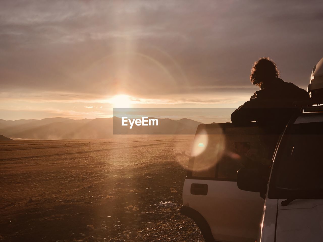 sky, sunset, nature, adult, travel, cloud, one person, sunlight, transportation, journey, mode of transportation, evening, vacation, trip, beauty in nature, back lit, holiday, silhouette, lens flare, landscape, sun, light, land, horizon, adventure, scenics - nature, dusk, environment, motor vehicle, men, road trip, leisure activity, car, mountain, outdoors, tranquility, women, young adult, sunbeam, sea, sitting, lifestyles, travel destinations, person, rear view, copy space, relaxation