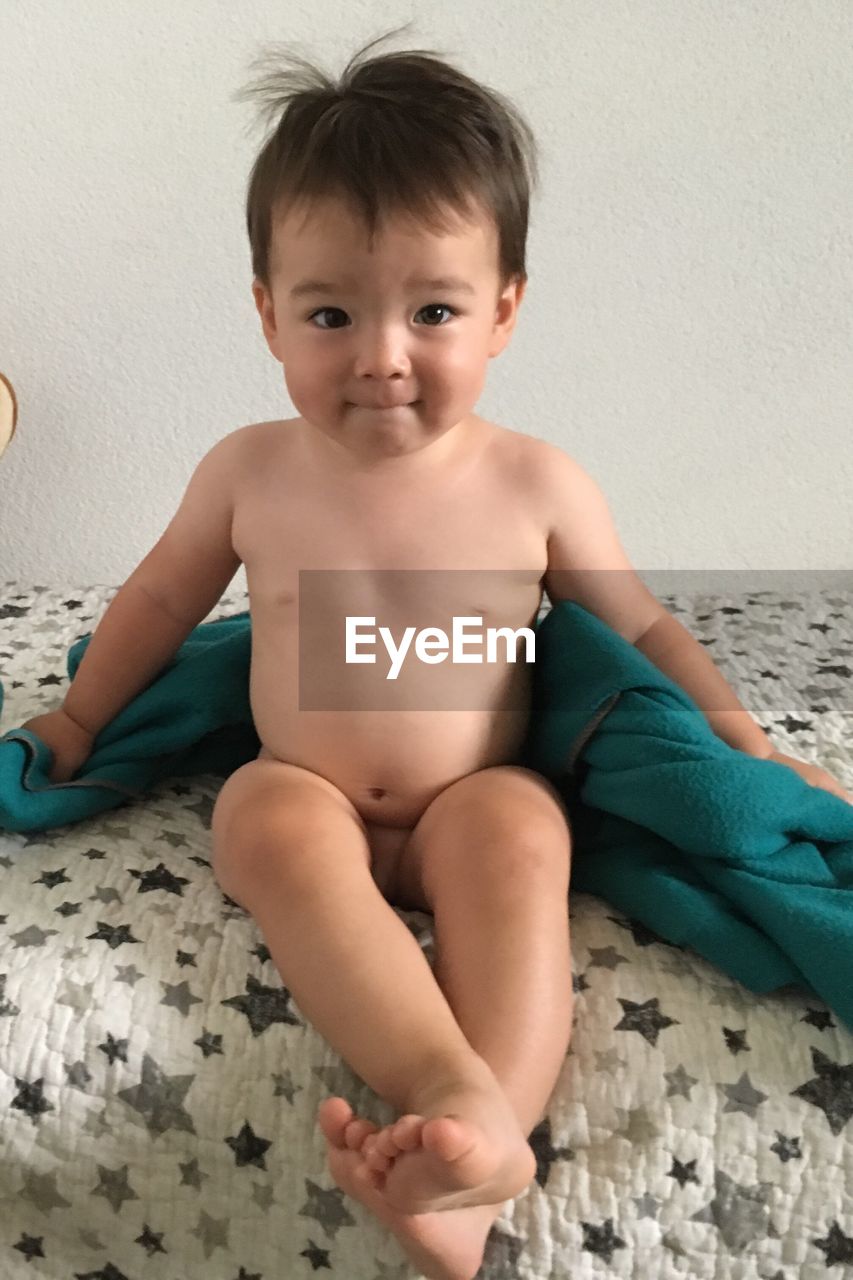 Portrait of naked baby boy with blanket sitting on bed