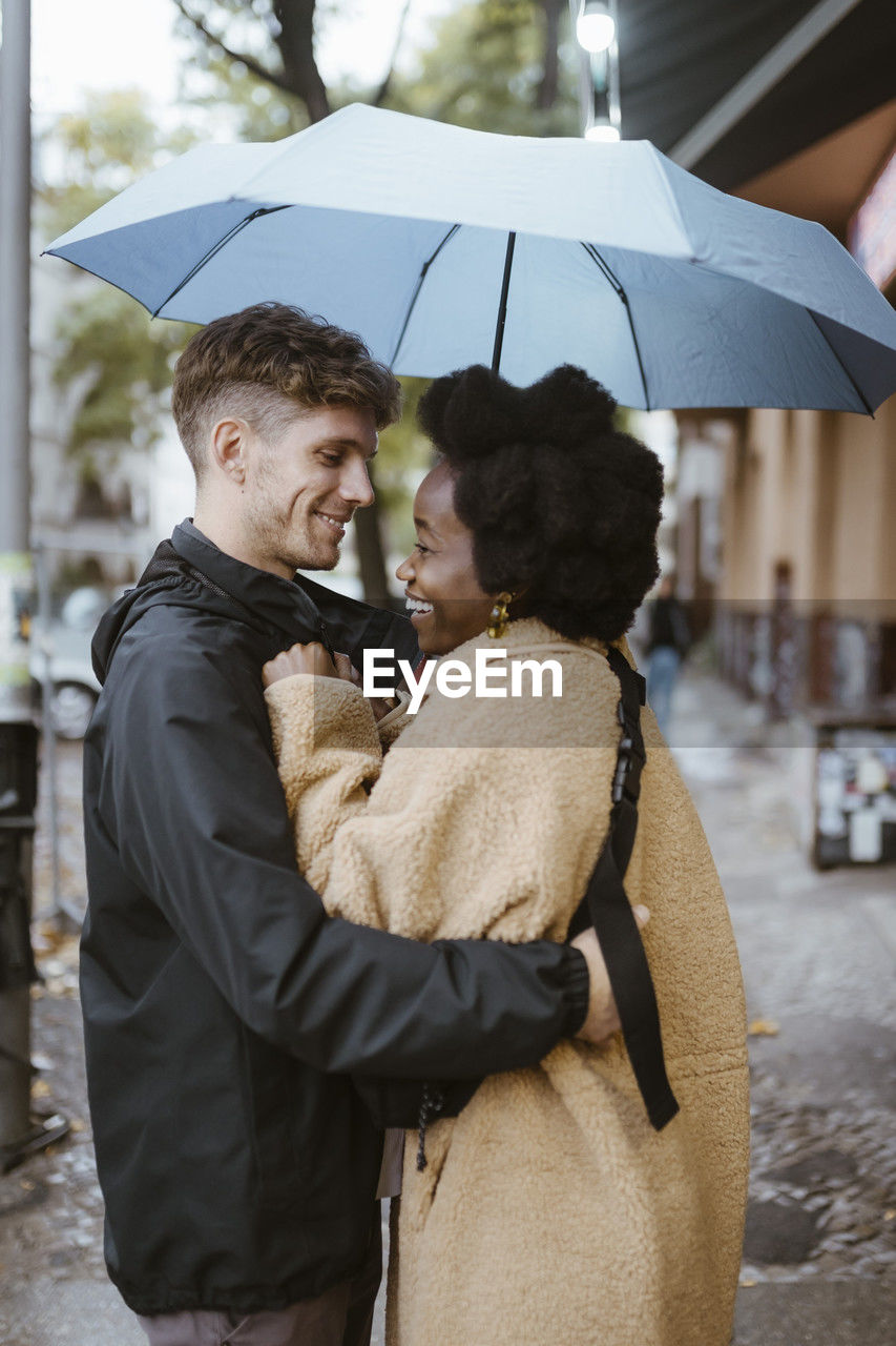 Side view of happy multiracial couple standing under umbrella at street