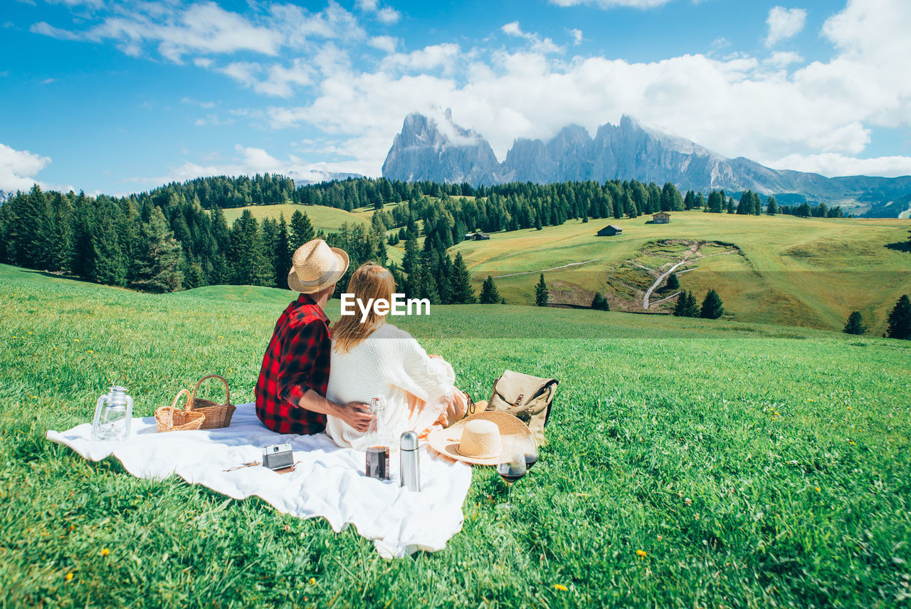 REAR VIEW OF COUPLE SITTING ON FIELD AGAINST MOUNTAINS