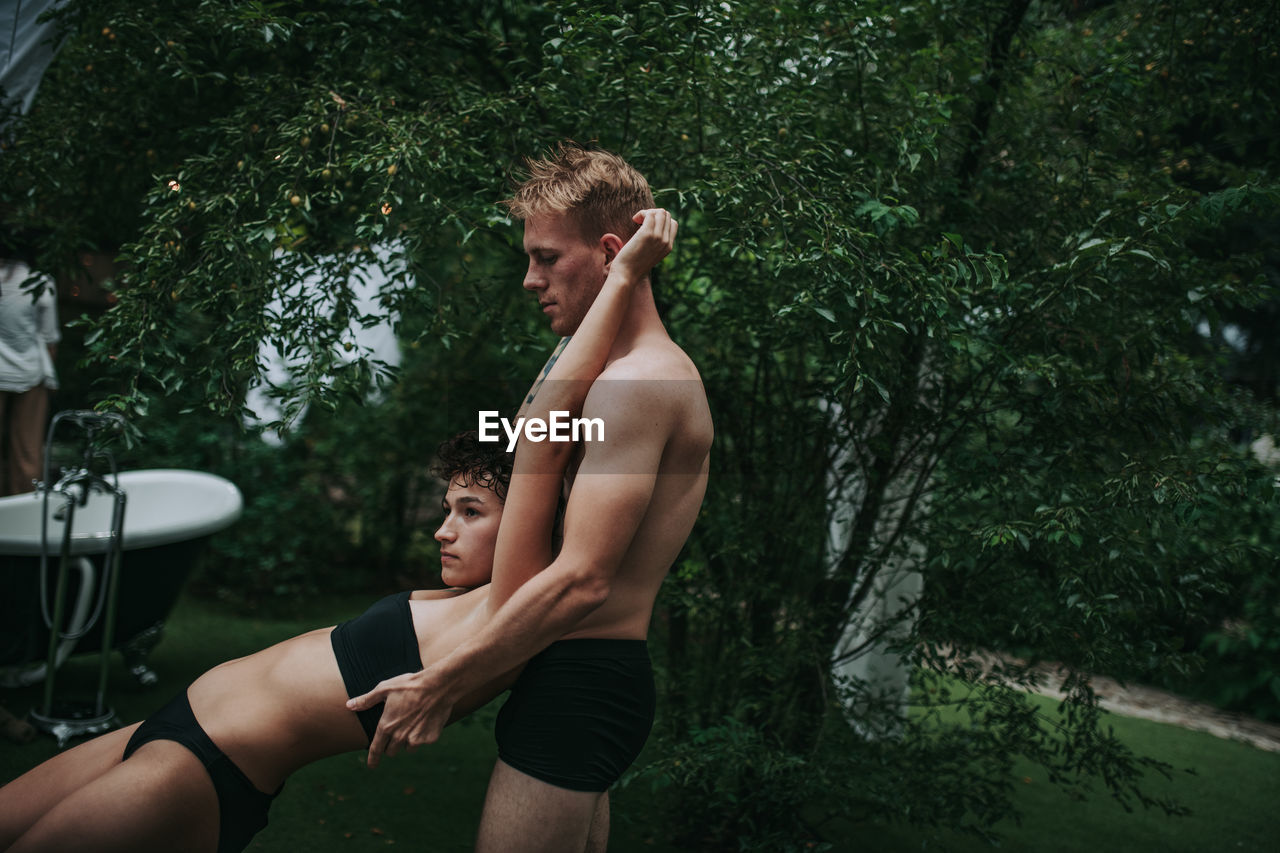 Side view of shirtless young man and plants against trees