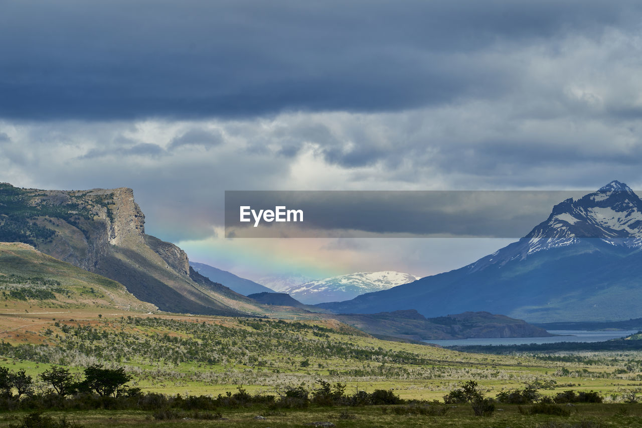 Vast open landscape in patagonia with dramatic sky and rainbow over a valley, snow covered mountains