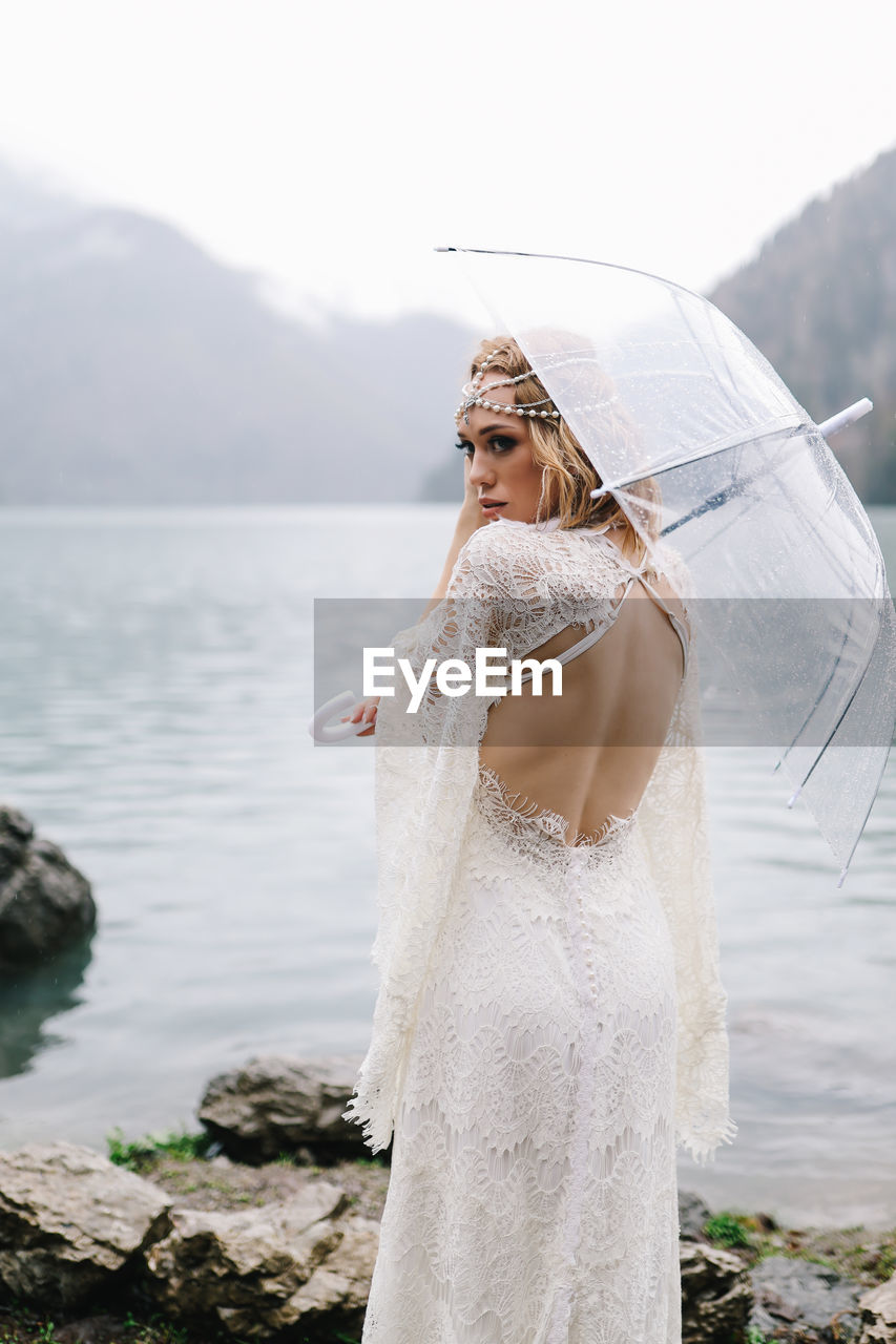 A beautiful young woman bride in a wedding lace dress stands in the middle of a lake and mountains