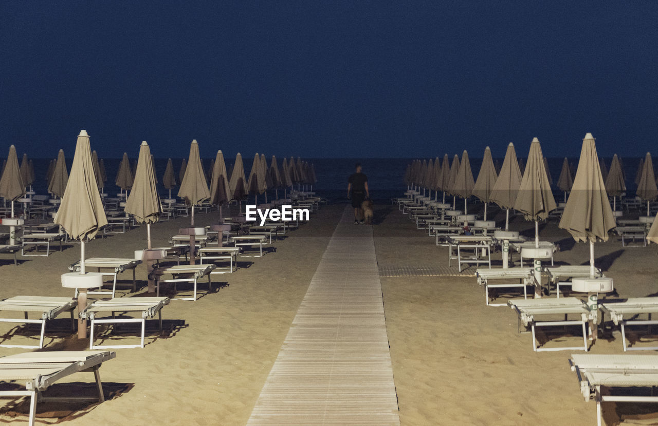 in a row, chair, sky, seat, nature, beach, land, sea, clear sky, arrangement, large group of objects, rear view, sand, architecture, table, water, travel destinations, outdoors, diminishing perspective, one person, adult, order, dock, sunlight