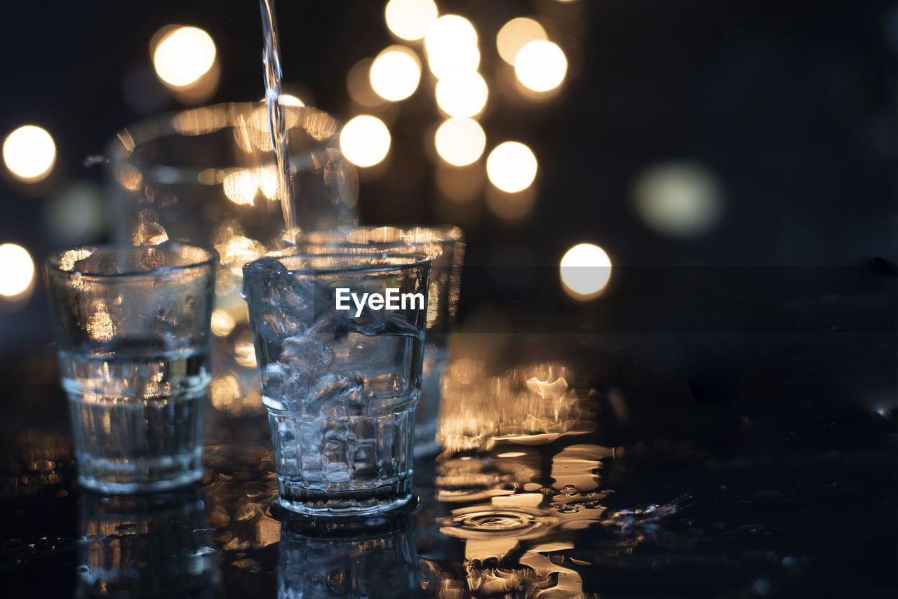 Shot glass, which is putting some clear liquid into the glass. the back is orange bokeh