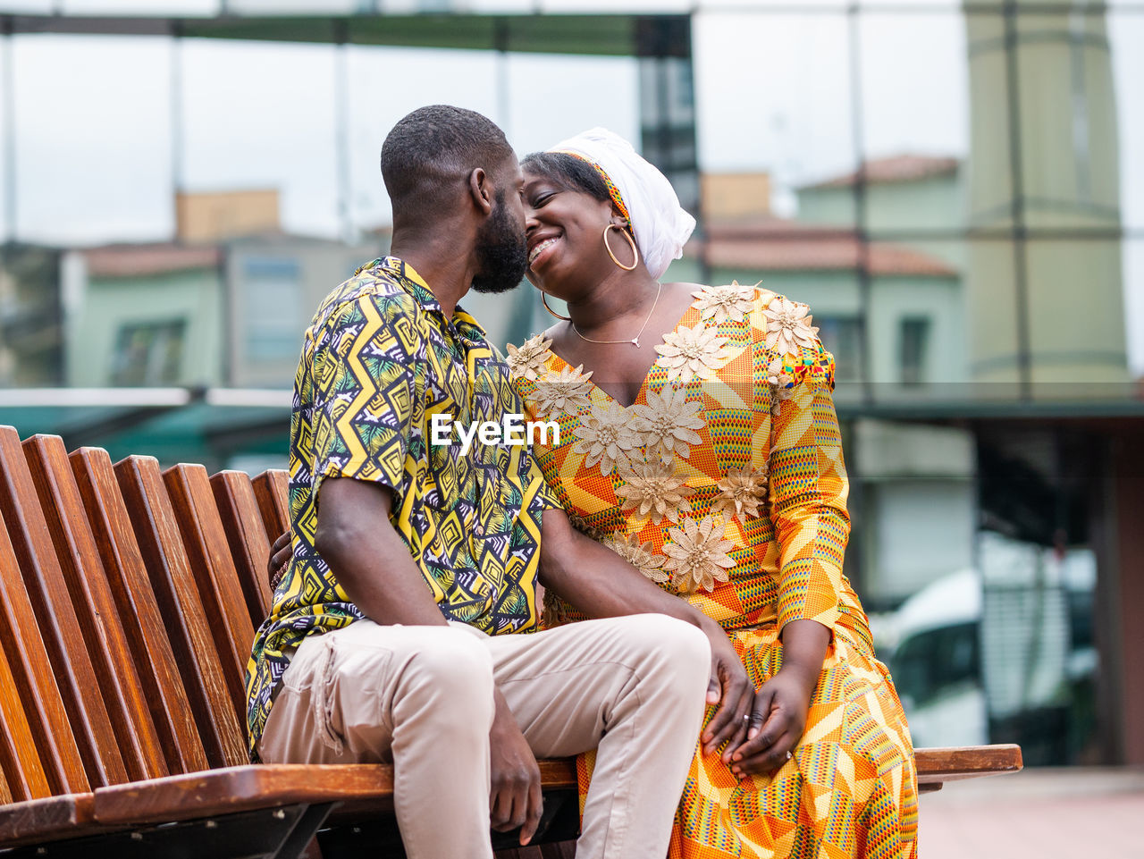 Cheerful african woman in traditional wear sitting close to unshaven boyfriend in ornamental clothes on city bench