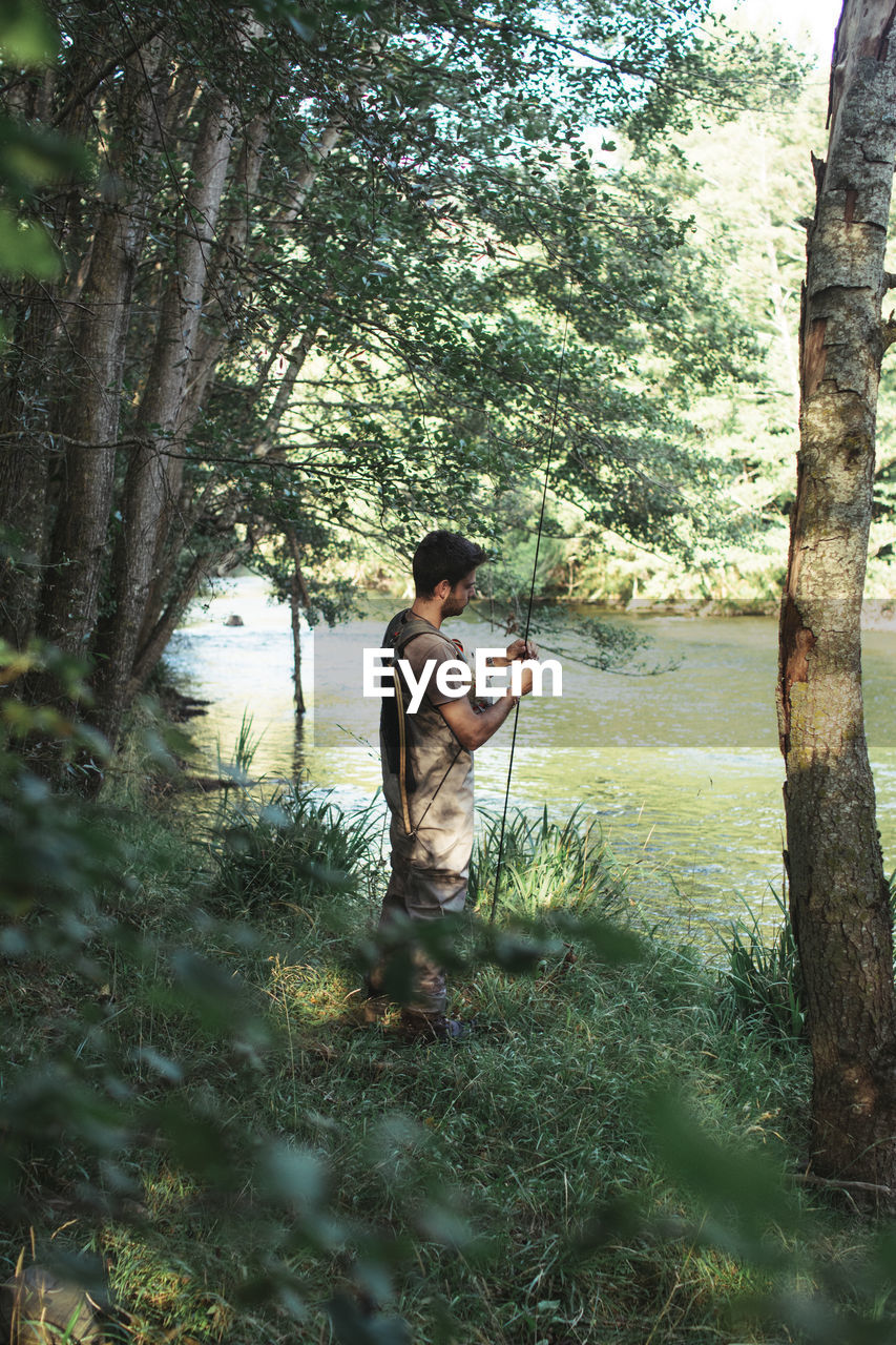 Side view of enthusiastic man preparing fishing rod into river in bright day