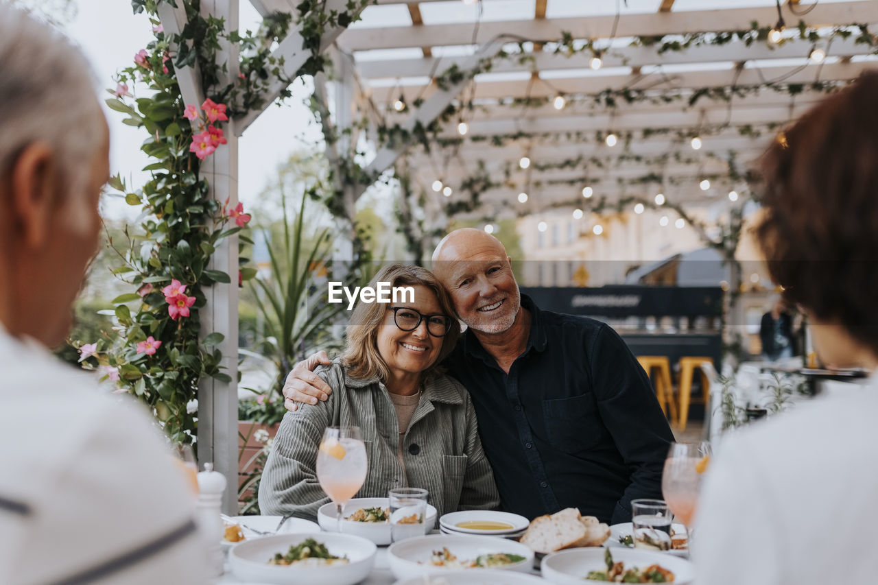 Portrait of smiling senior man with arm around elderly female friend sitting at restaurant