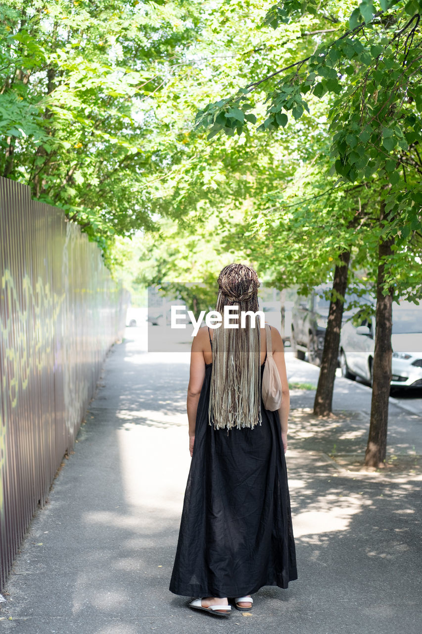 one person, tree, plant, full length, dress, spring, rear view, adult, nature, women, day, clothing, walking, outdoors, footpath, fashion, city, lifestyles, young adult, hairstyle, sunlight, standing, green, leisure activity, architecture, road