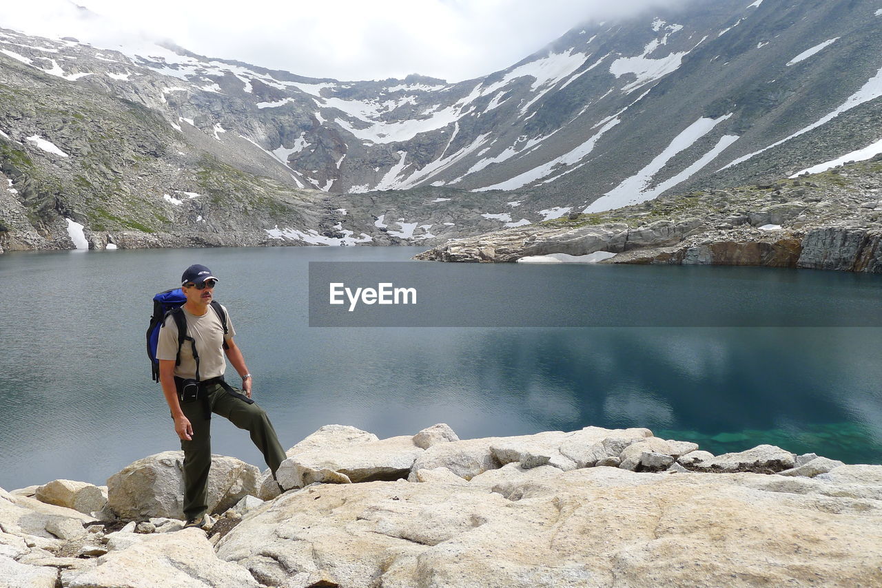 Hiker standing on rock at lakeshore against snow covered mountain