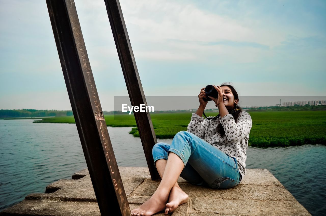 Young woman photographing through camera while sitting against lake