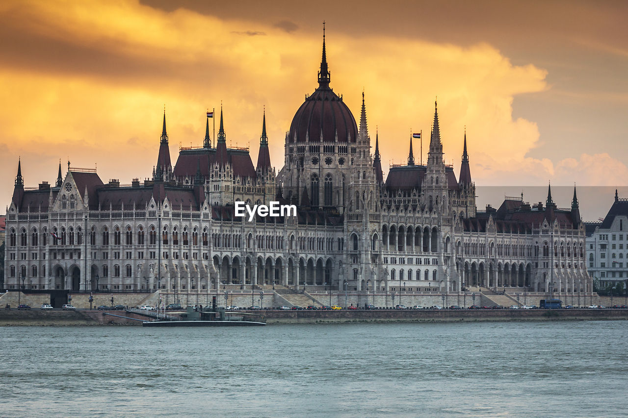 Hungarian parliament building by river against cloudy sky