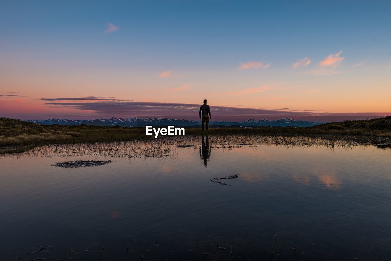 Rear view of man standing by lake against sky during sunset