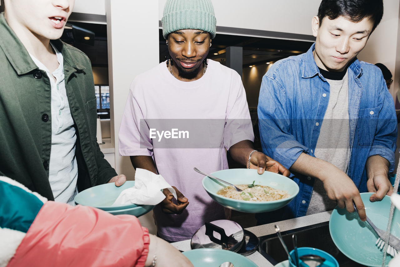 Young man with noodles while roommate cleaning dishes at sink in college dorm