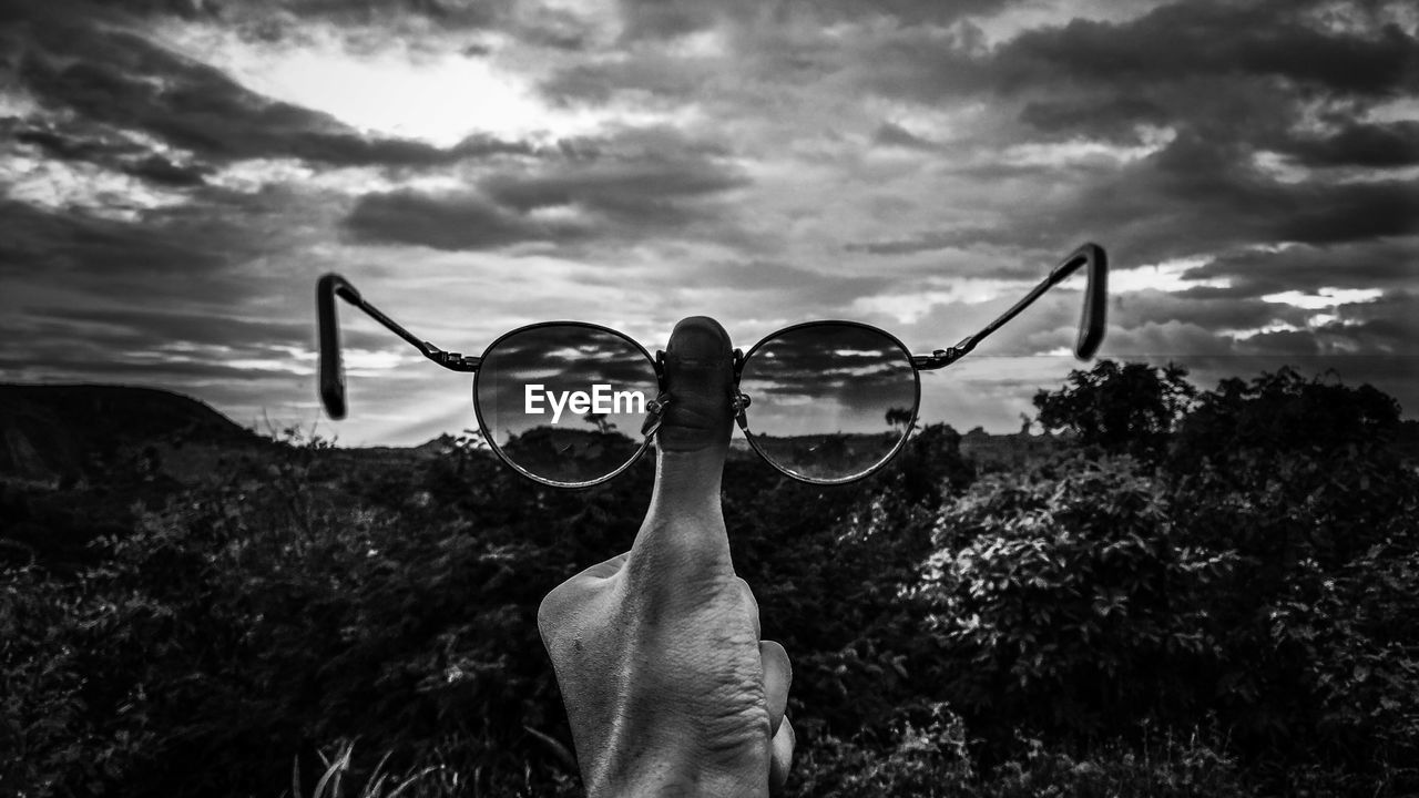 Cropped hand holding sunglasses by trees against cloudy sky