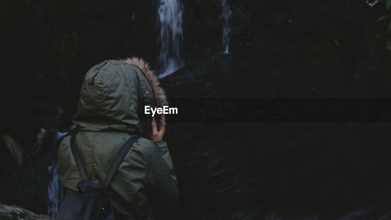 Rear view of woman in warm clothing standing against waterfall