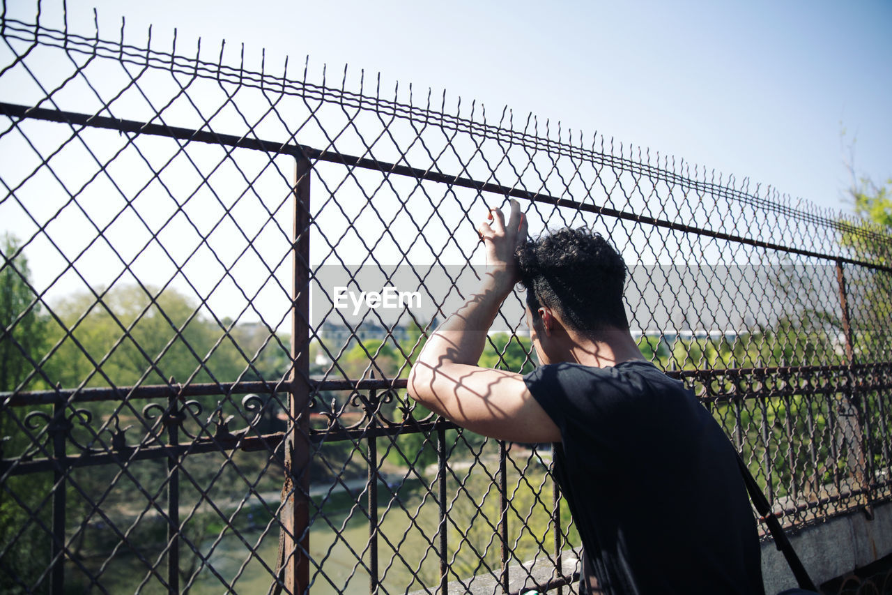 Man looking through chainlink fence against sky