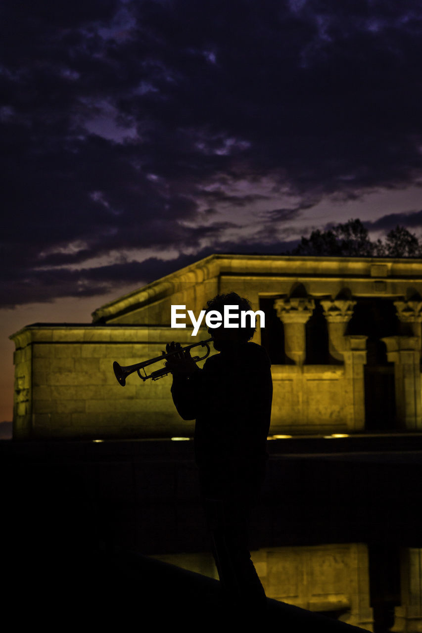 Silhouette man playing trumpet against temple of debod at night