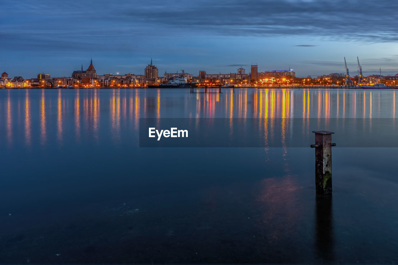 Illuminated buildings by sea against sky at dusk