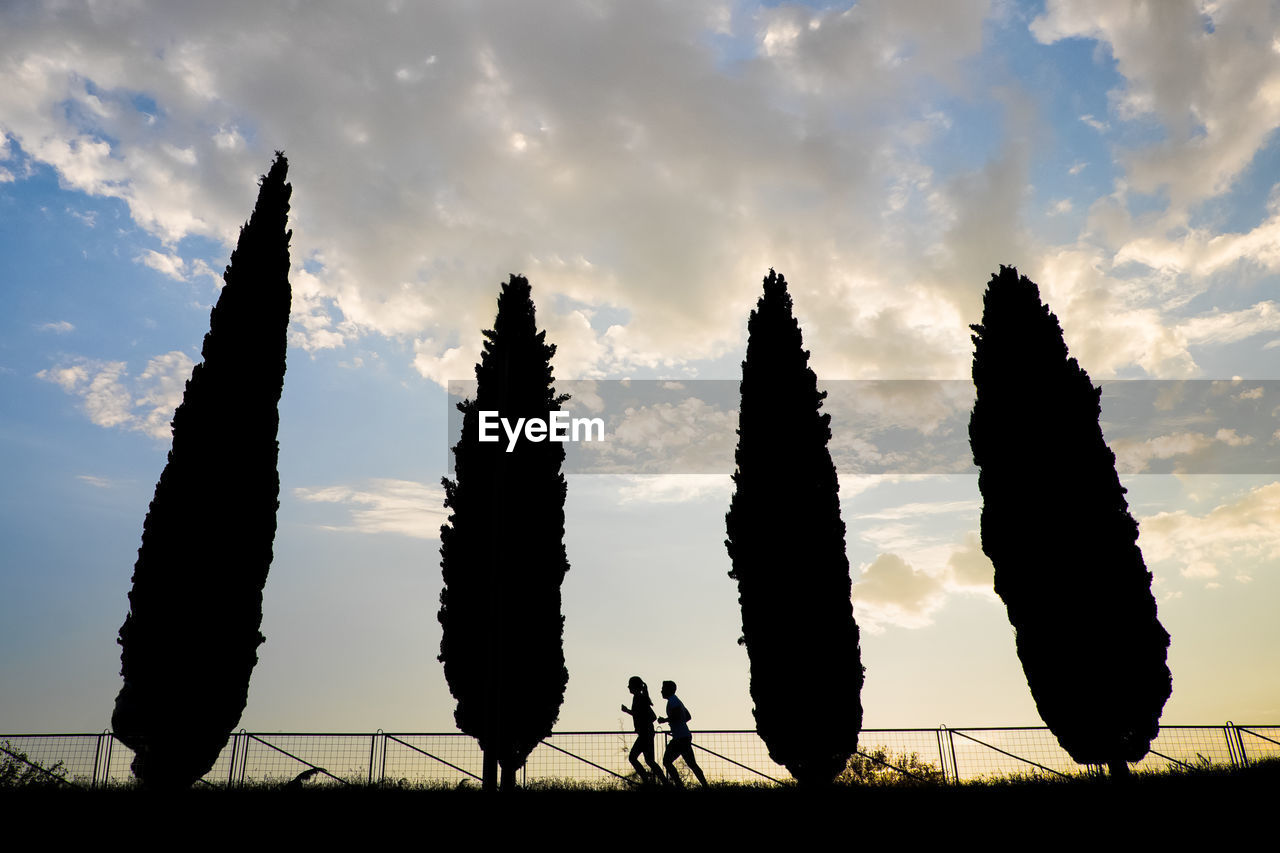 Low angle view of silhouette couple jogging by trees against cloudy sky during sunset