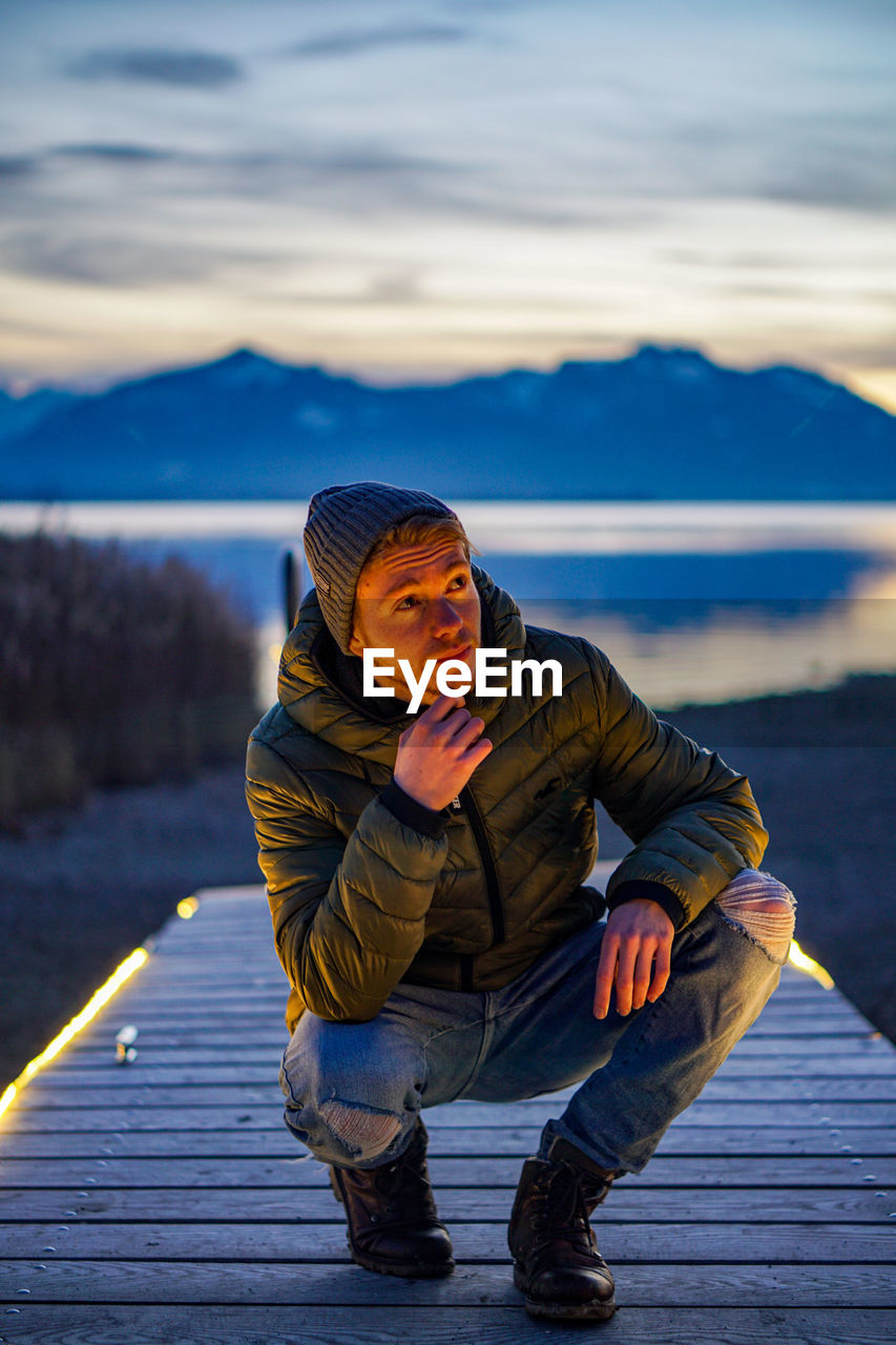 Full length of young man crouching on boardwalk against sky during sunset