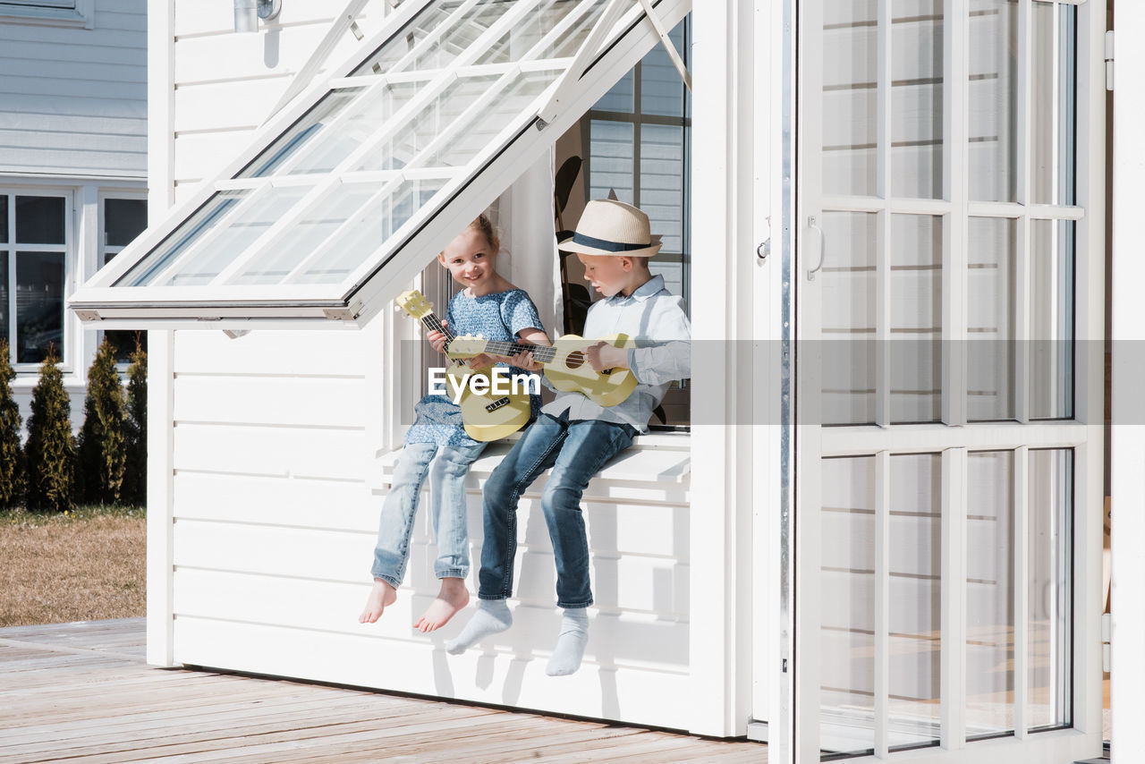 Brother and sister sat in a window at home playing guitar together