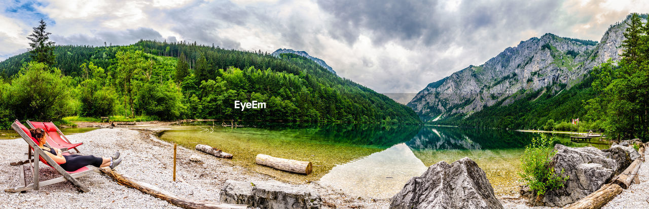 Panoramic view of woman sitting on lounge chair by lake and mountains against sky