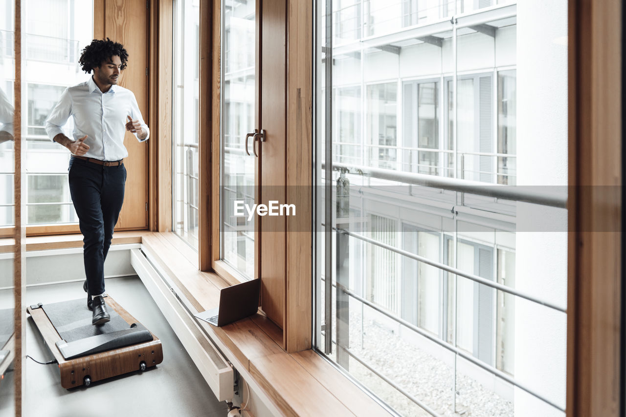 Active male entrepreneur walking on treadmill while looking at laptop against window in workplace