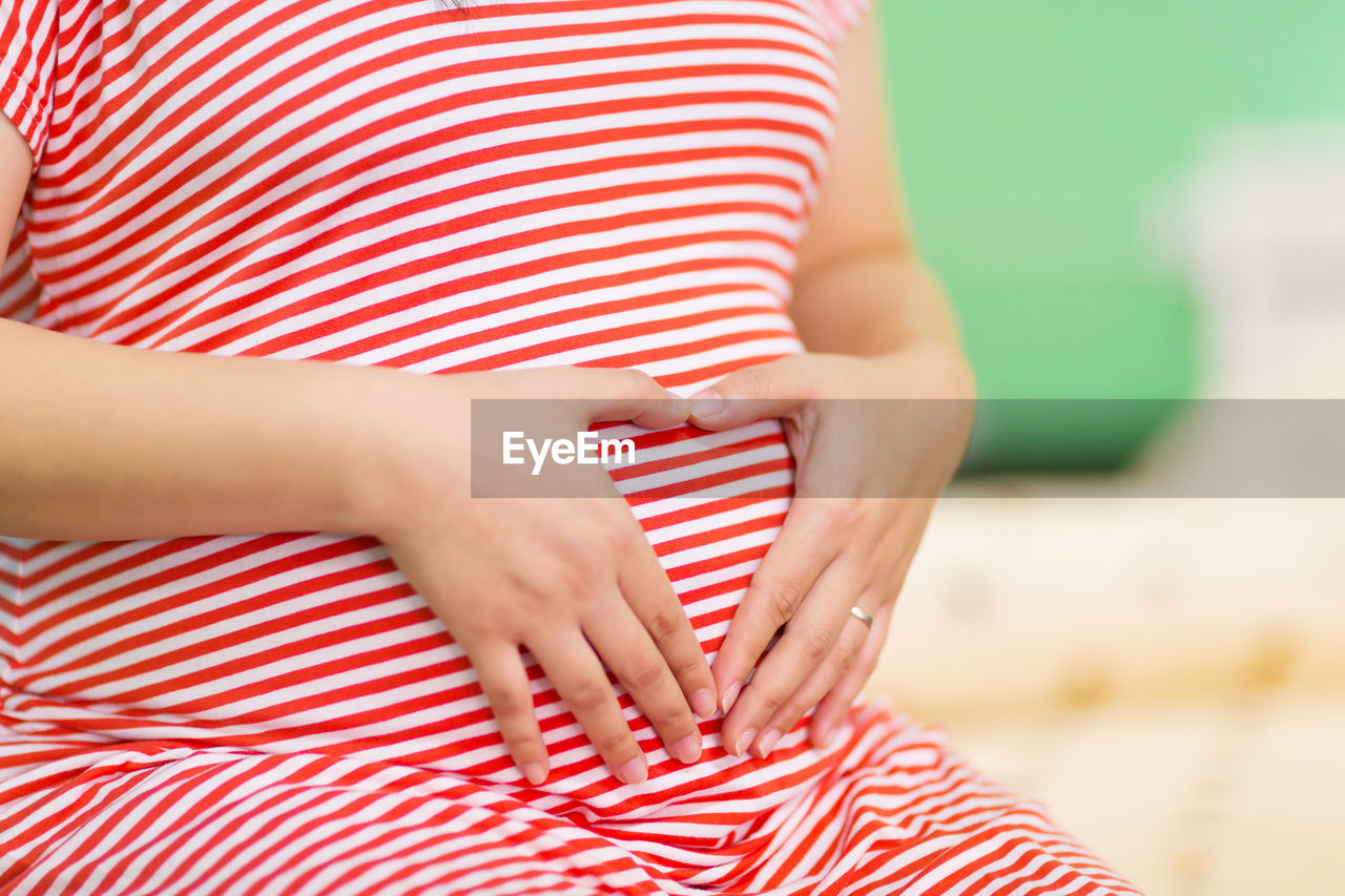 Midsection of pregnant woman making heart shape on abdomen indoor